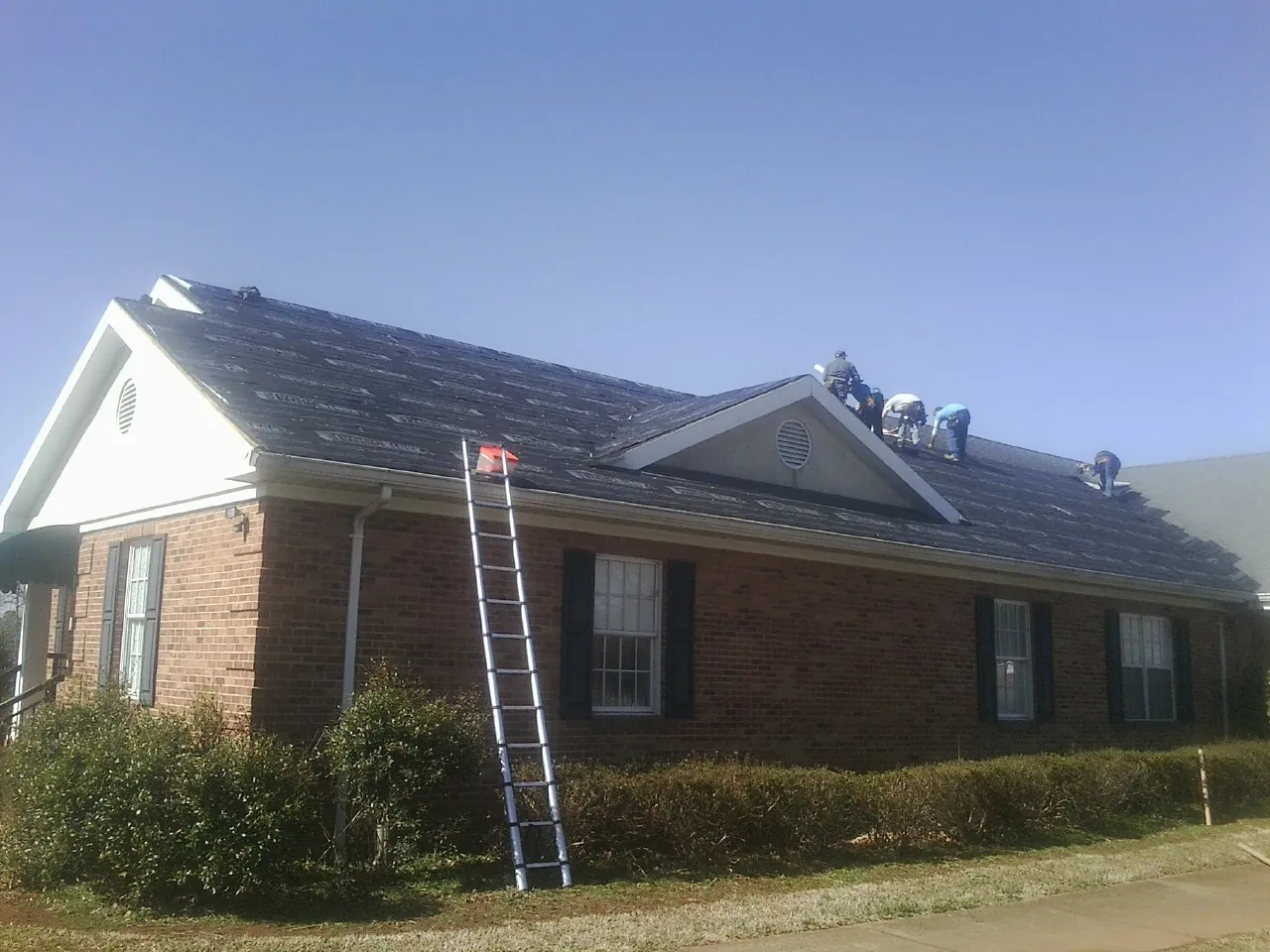 Crew tearing shingles off a brick ranch roof with a ladder set — Metal Roof Repair work by our crew in Metamora
