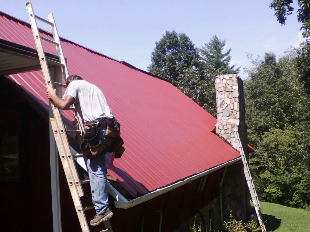 Roofer on a ladder beside a red metal roof and stone chimney — Soffit & Fascia Repair by Roofing Brothers in Metamora